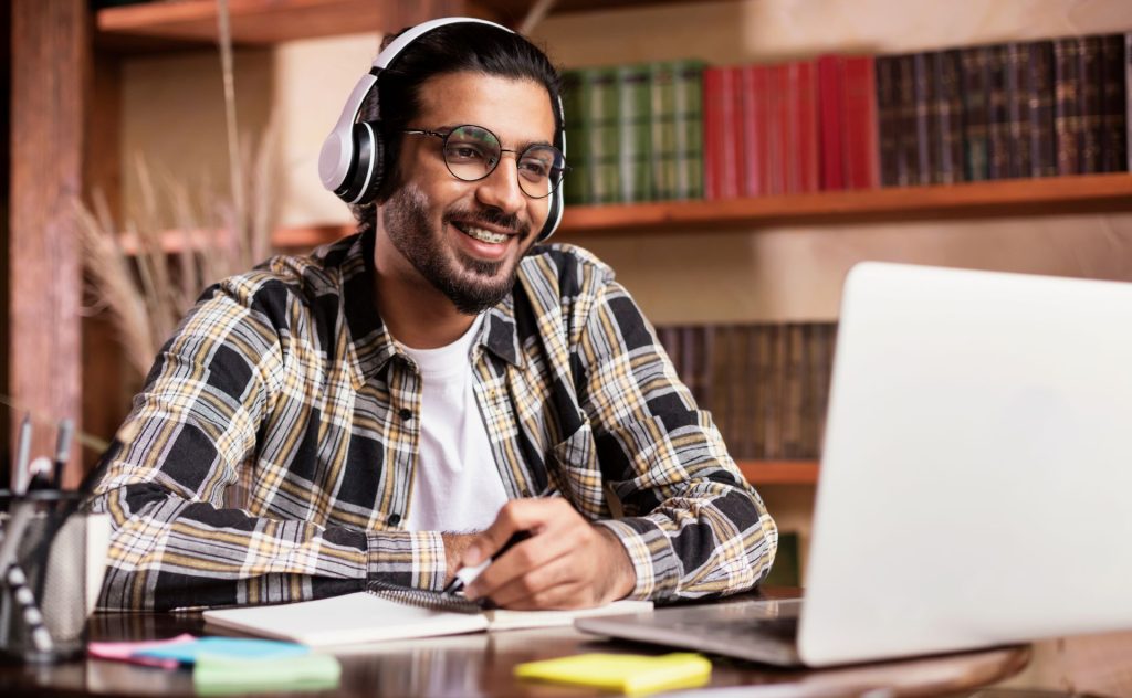 A man with headphones on is smiling, holding a pen above a notebook and looking into a computer. He is in a library or office.