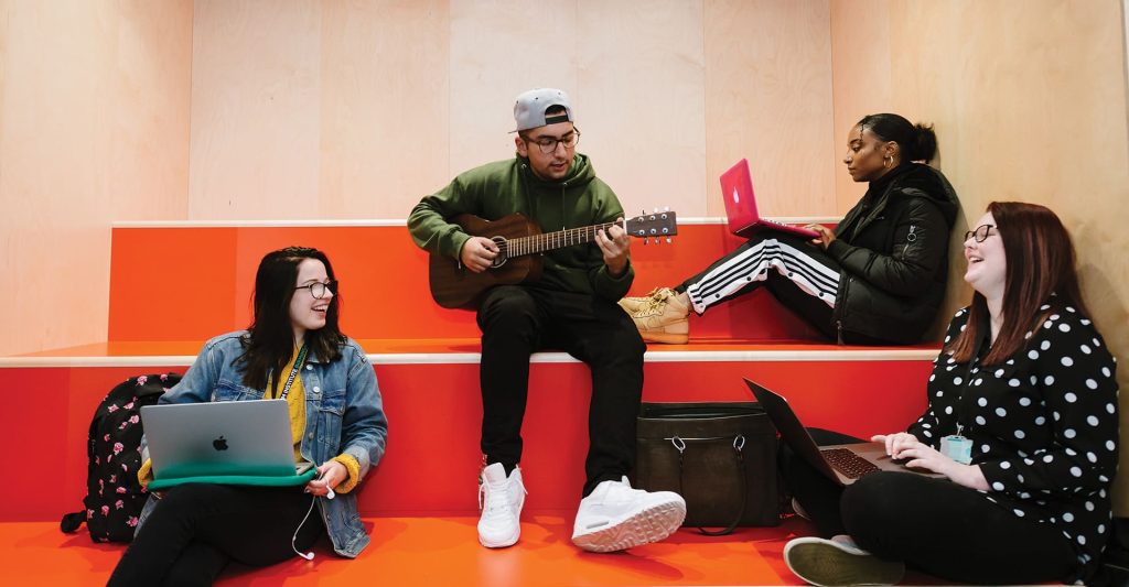 Four students sitting on red stairs, one is in the middle playing a guitar. The other three are sitting around them - the other three on their laptops. Everyone is smiling.